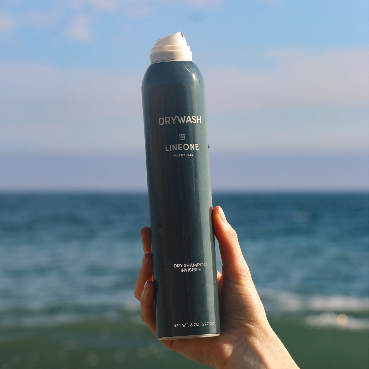Hand holding a can of DRYWASH invisible dry shampoo by LINEONE Hair against the ocean