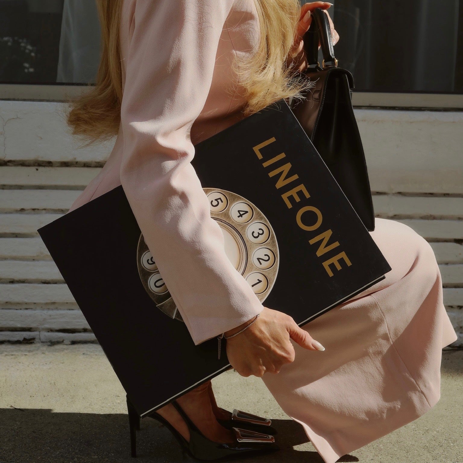 Stylist in pink suit holding LINEONE Hair Bookcase volume with gold rotary-dial cover on steps