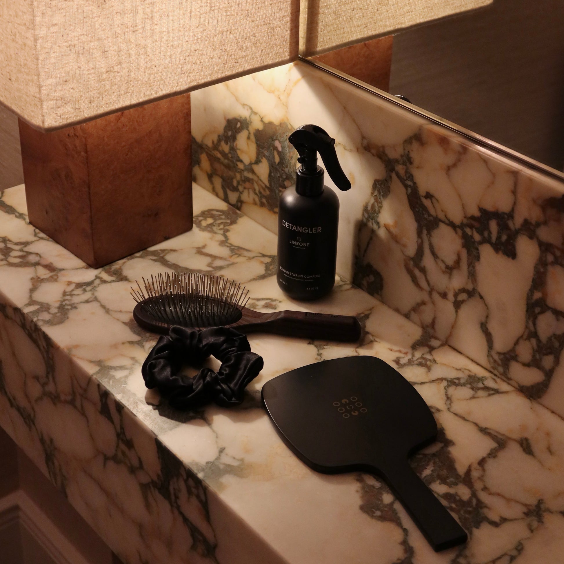 LINEONE Hair Mirror beside paddle brush, black silk scrunchie and Detangler under a linen lamp on marble