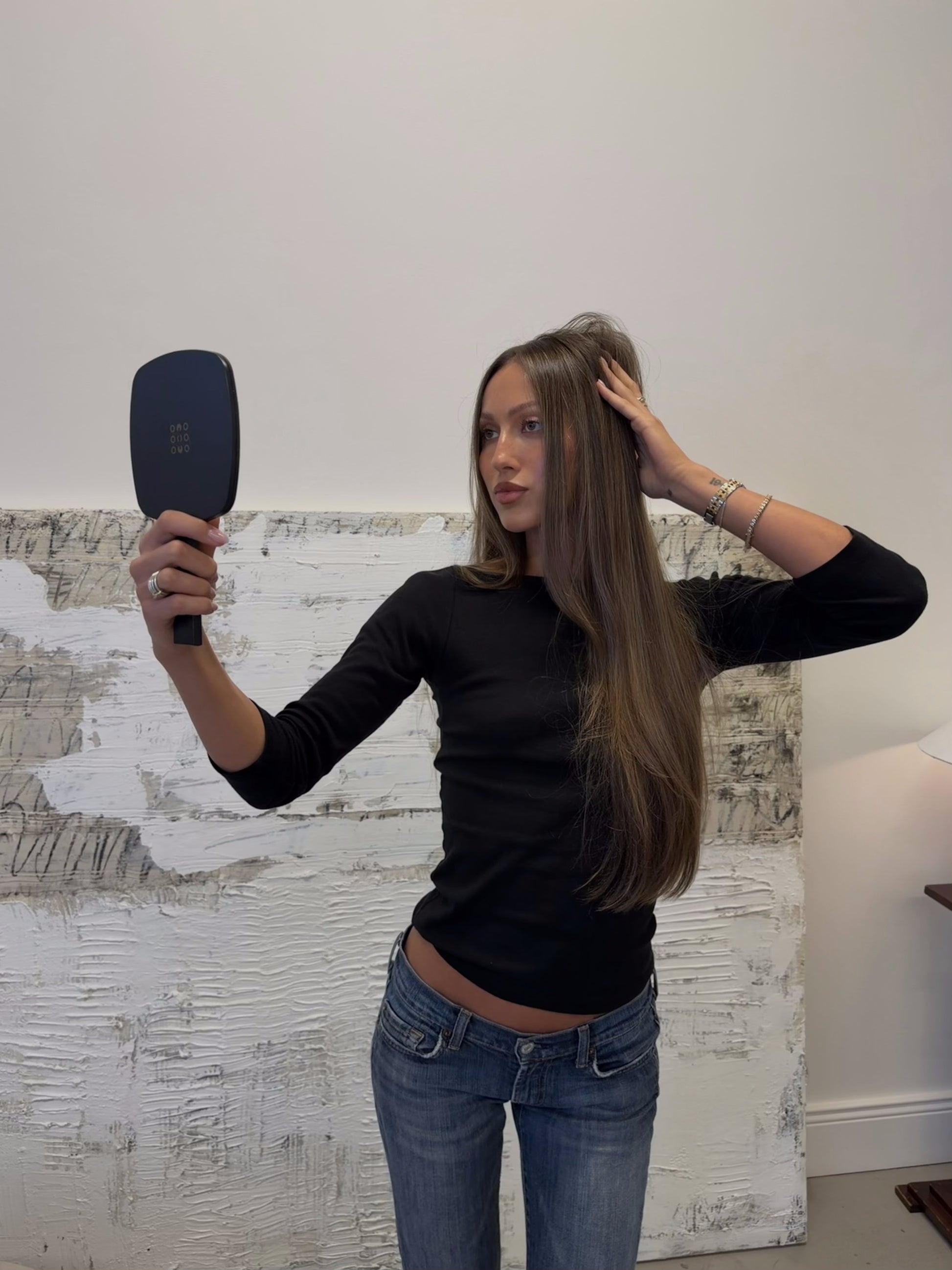 Model in black long-sleeve and jeans holding the LINEONE Hair Mirror up to her long brunette waves