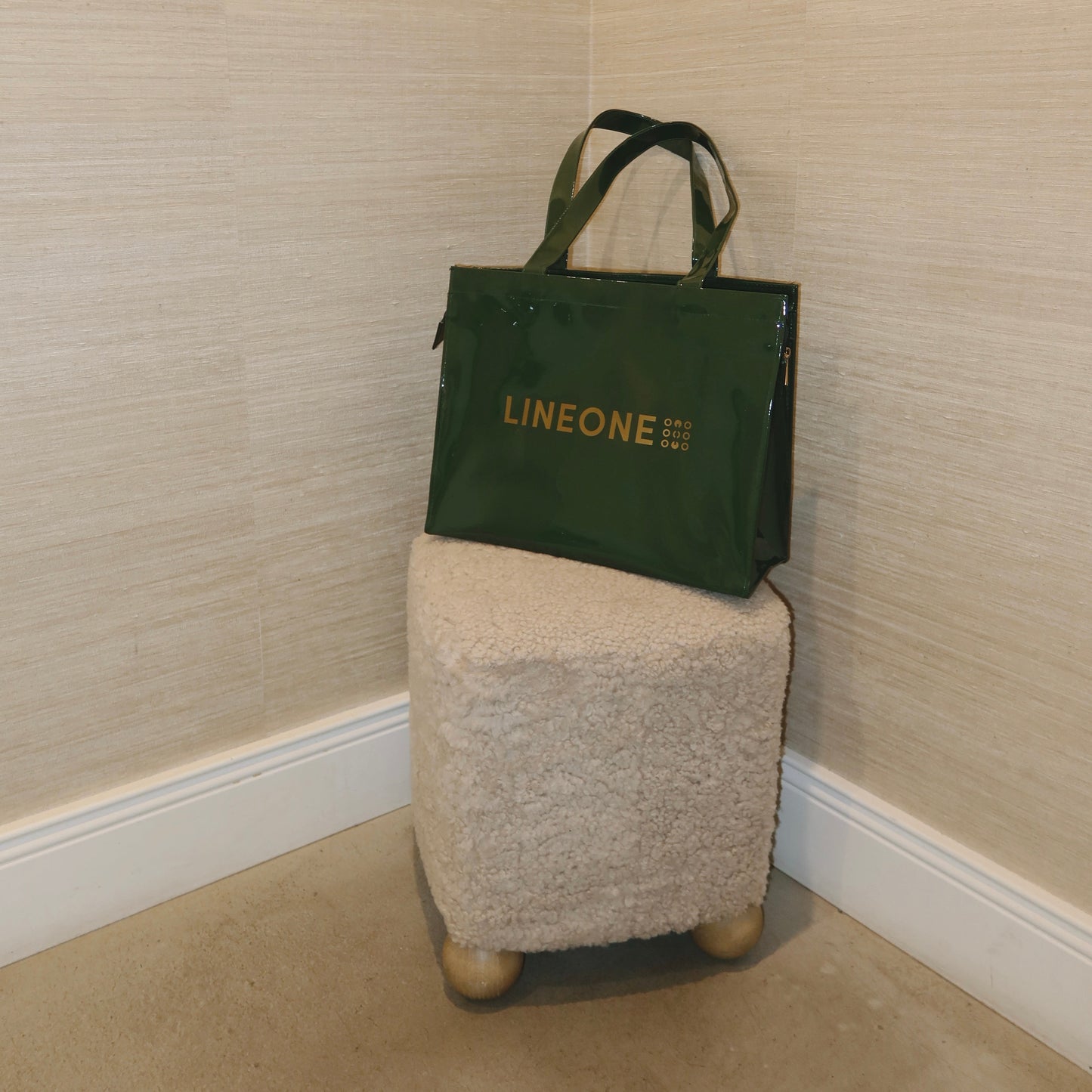 Hunter green LINEONE Hair Rodeo Bag perched on a small boucle ottoman in a corner with grasscloth walls